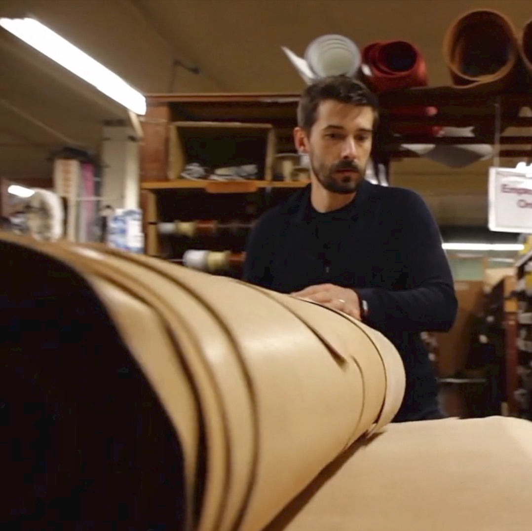 A man unrolling a hide of leather across a large table in a cluttered leather supply store