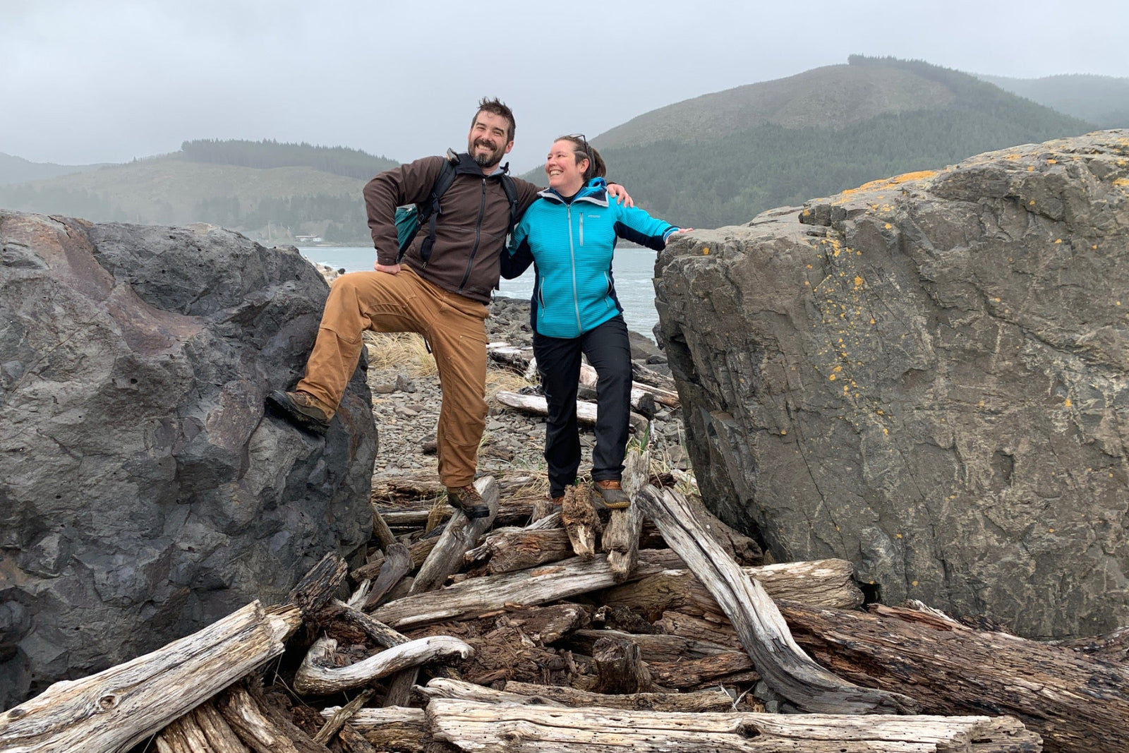 A man and a woman on a pile of driftwood on a misty , rainy Oregon beach with mountains and boulders