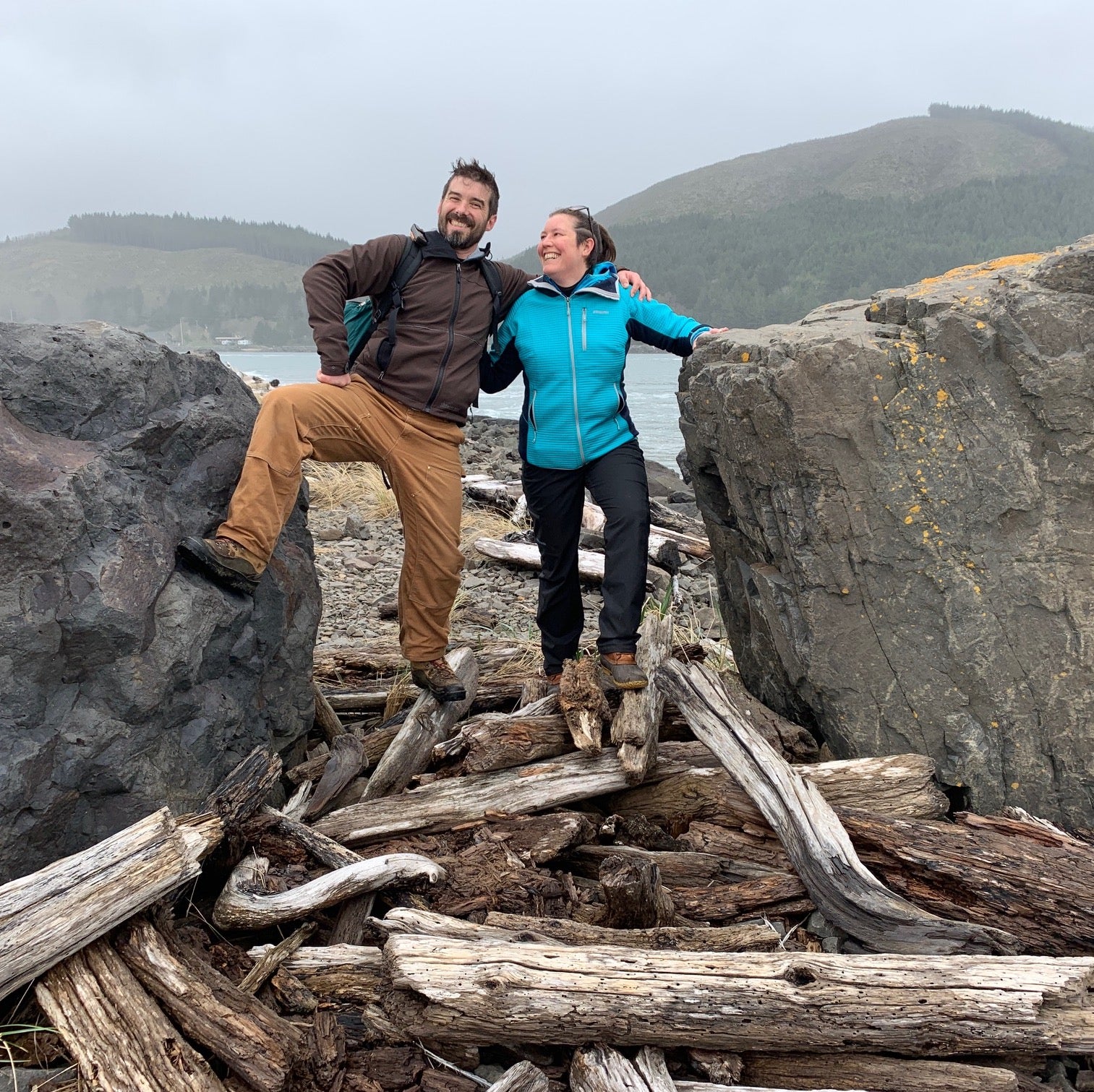 Valerie and Geoff from Walnut Studiolo are standing on a pile of driftwood between two boulders on the North Oregon Coast beach near Manzanita, with mountains and grey sky in the background