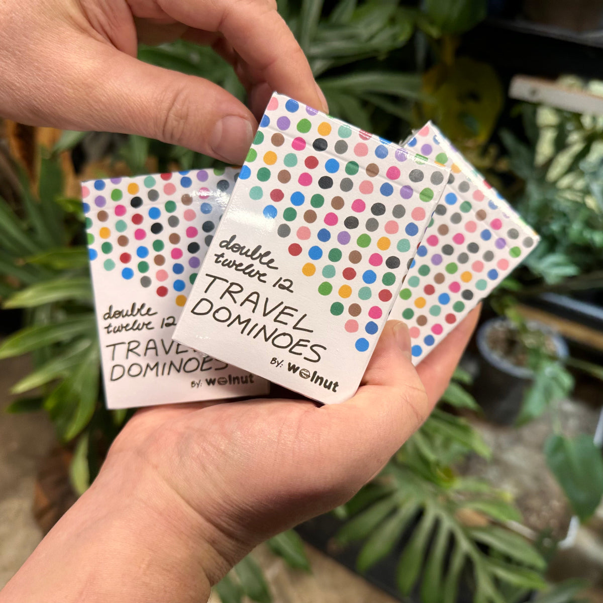 Two hands holding three packs of Walnut Travel Dominoes, featuring a colorful polka-dot pattern on the packaging, with a blurred green plant background.