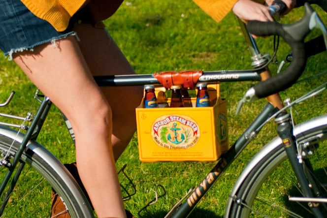 A woman bicycling on a bright summer day over a green lawn wearing suede fringe cowgirl boots and with a 6-pack of Anchor Steam beer connected to the top tube of her bicycle with a honey leather 6-pack frame cinch