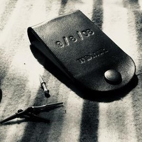 A black and white, dramatic, noir photo of a closed pocket-sized leather travel cribbage board on a striped blanket, which is monogrammed with a couple's wedding anniversary date, 9/8/08.