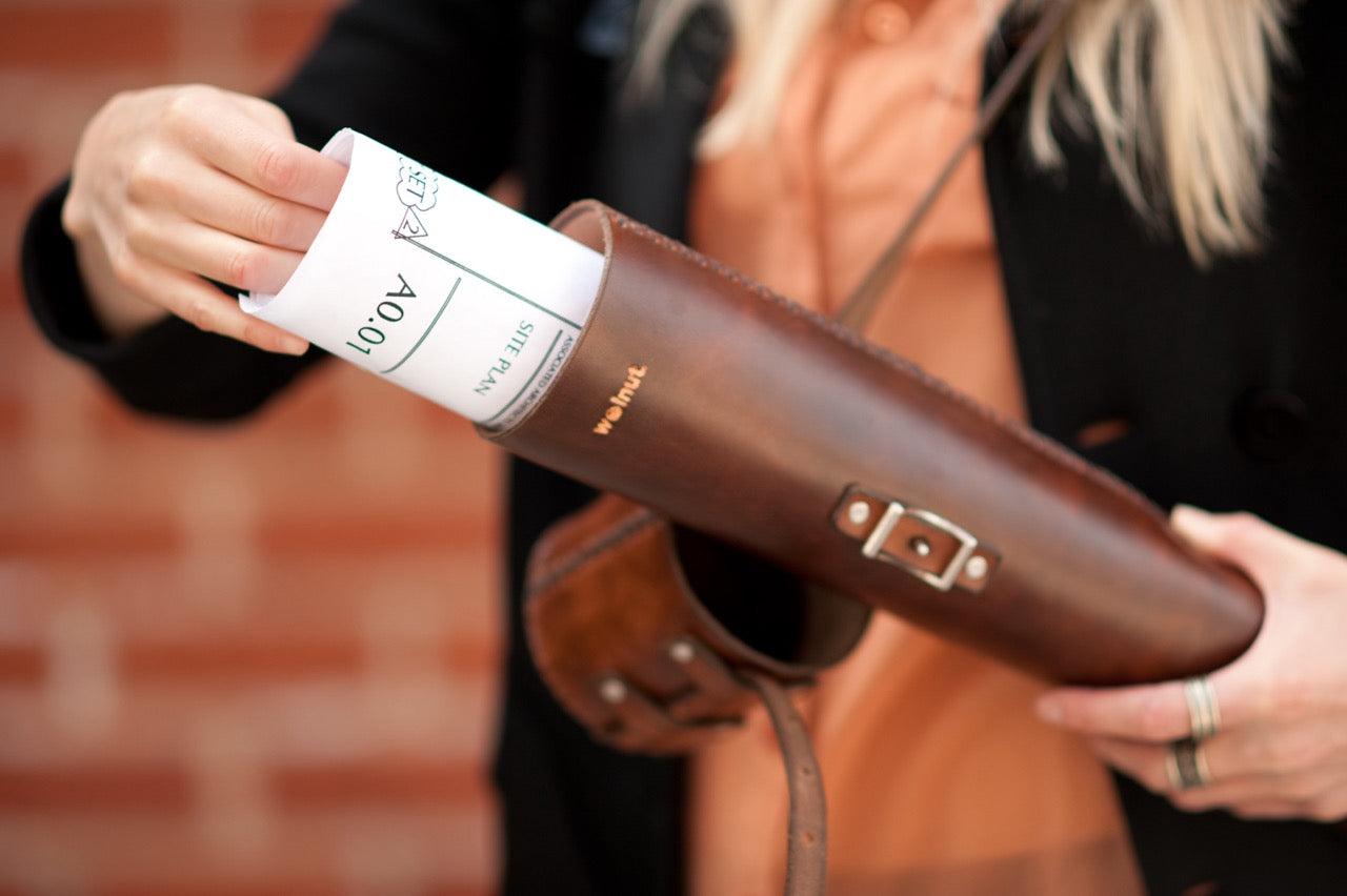 A woman in an orange shirt and black cardigan is pulling a set of architectural plans out of a hand-stitched leather storage tube in front of a brick wall