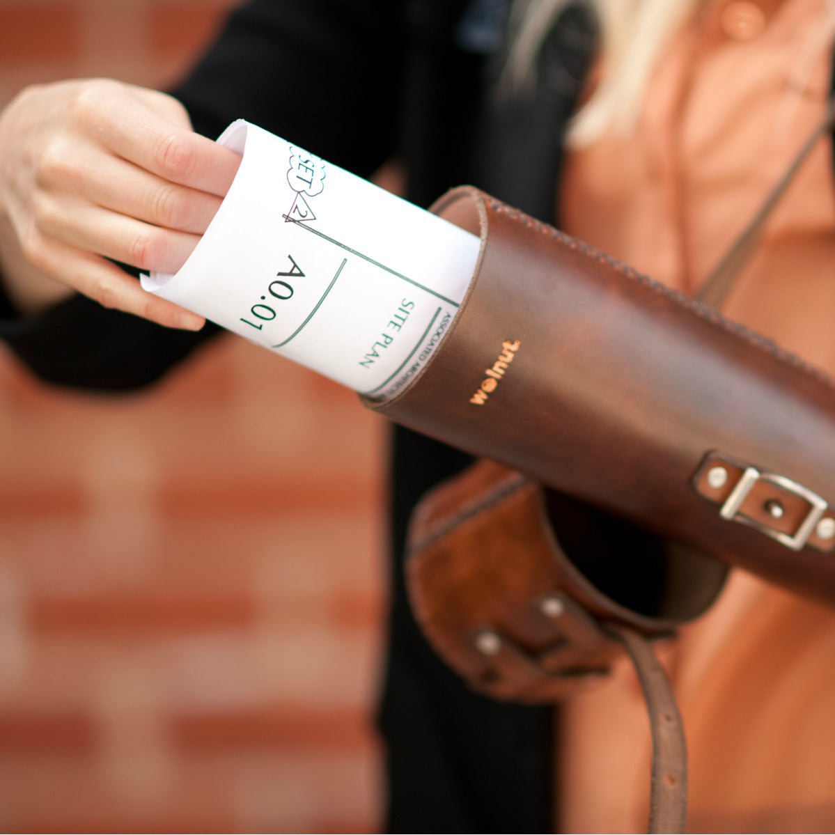 A woman in an orange shirt and black cardigan is pulling a set of architectural plans out of a hand-stitched leather storage tube in front of a brick wall