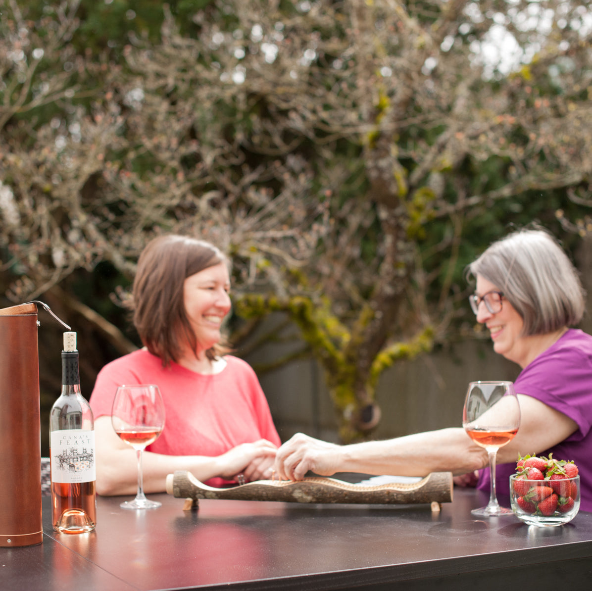 A young woman and her mother playing a game of cribbage on a natural branch wood board with a bottle of rose wine and a handcrafted leather wine bottle case