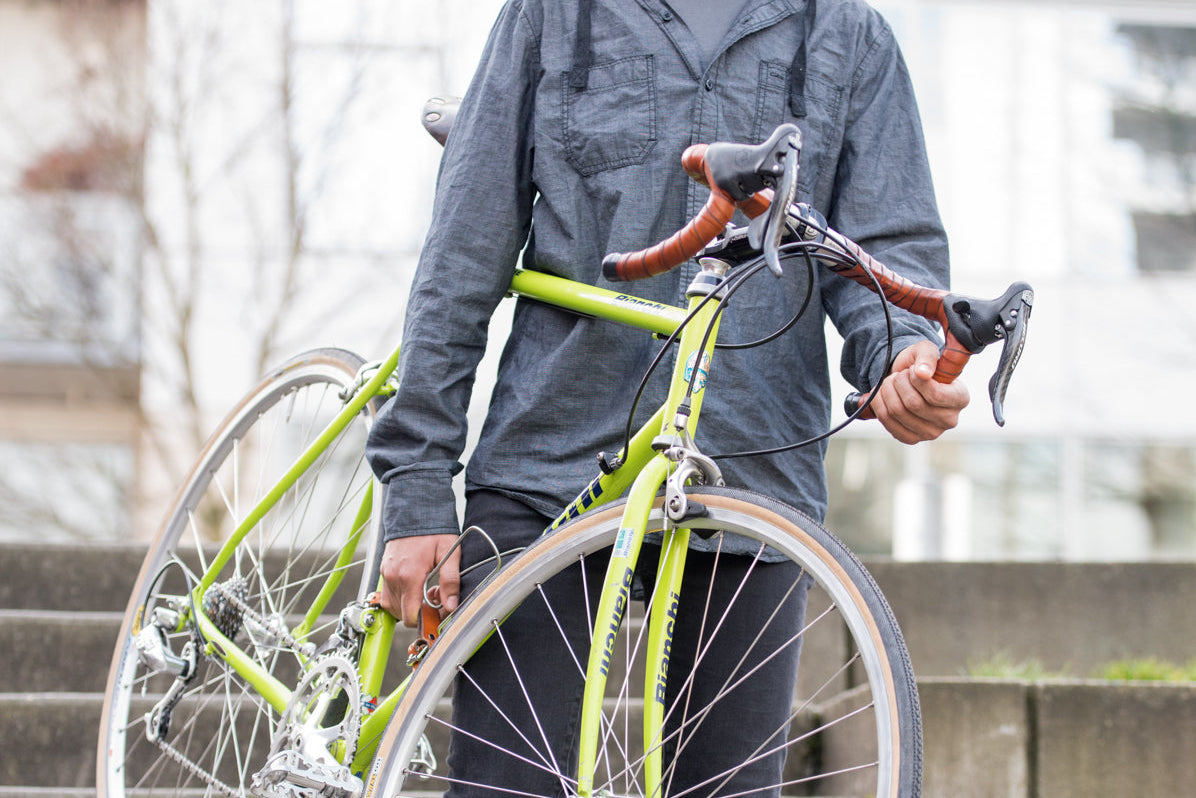 A Black woman wearing a denim shirt is carrying a bicycle down a set of stairs in an urban environment. The bicycle has a green paint job and honey leather wrapped handlebars.
