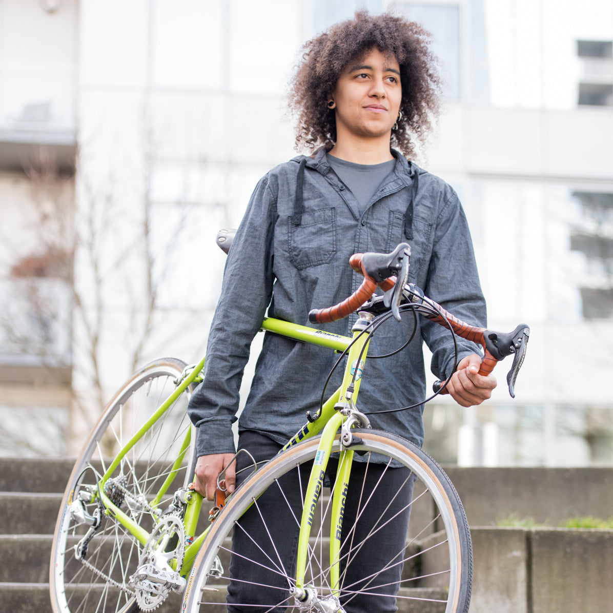 A woman carrying a bicycle down a set of stairs in a city park in front of an urban building. The bicycle has a leather frame carrying strap and leather handlebar wraps.