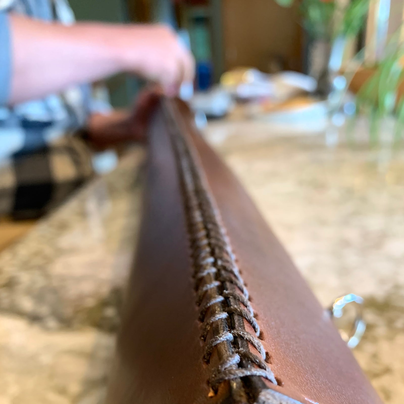 A long dark brown leather blueprint tube is shown on a long countertop being hand stitched together by Geoff, whose stitching hands are in the background