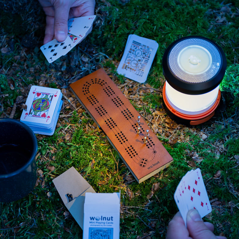 Small folding leather cribbage board being played at night while camping with an LED lantern and half-size mini playing cards for backpacking.