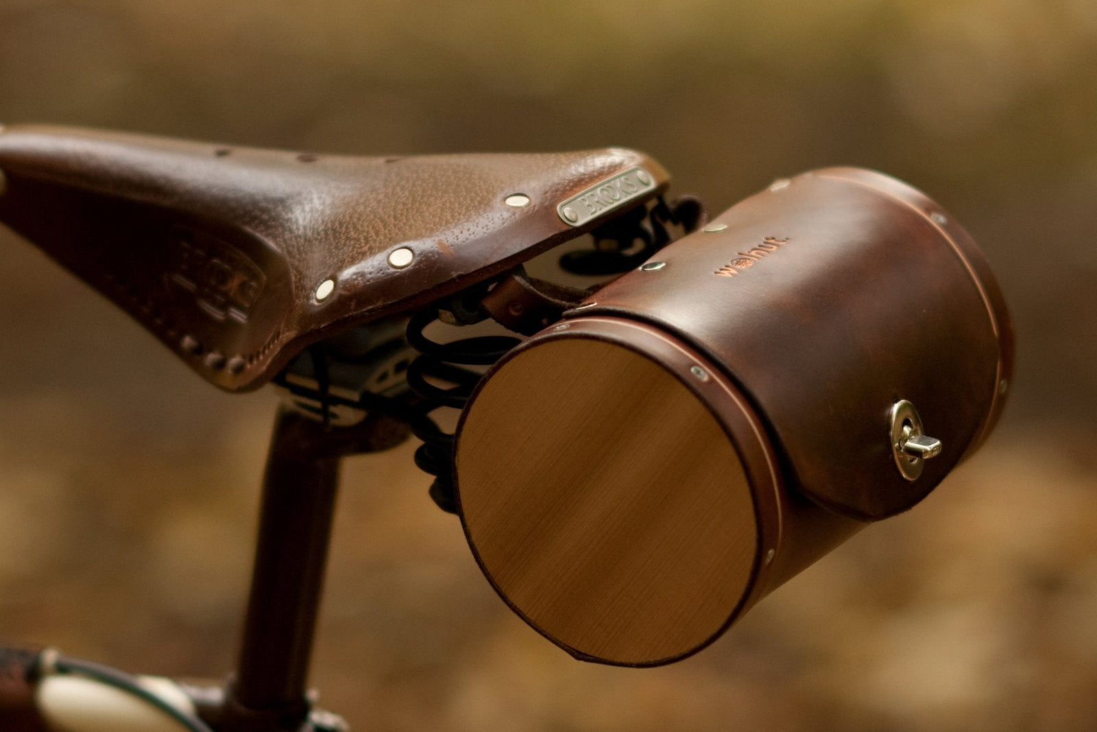 A dark brown leather round, barrel-shaped saddle bag with cedar wood sides mounted to the back of a brown leather Brooks saddle on a cream-and-brown painted bicycle