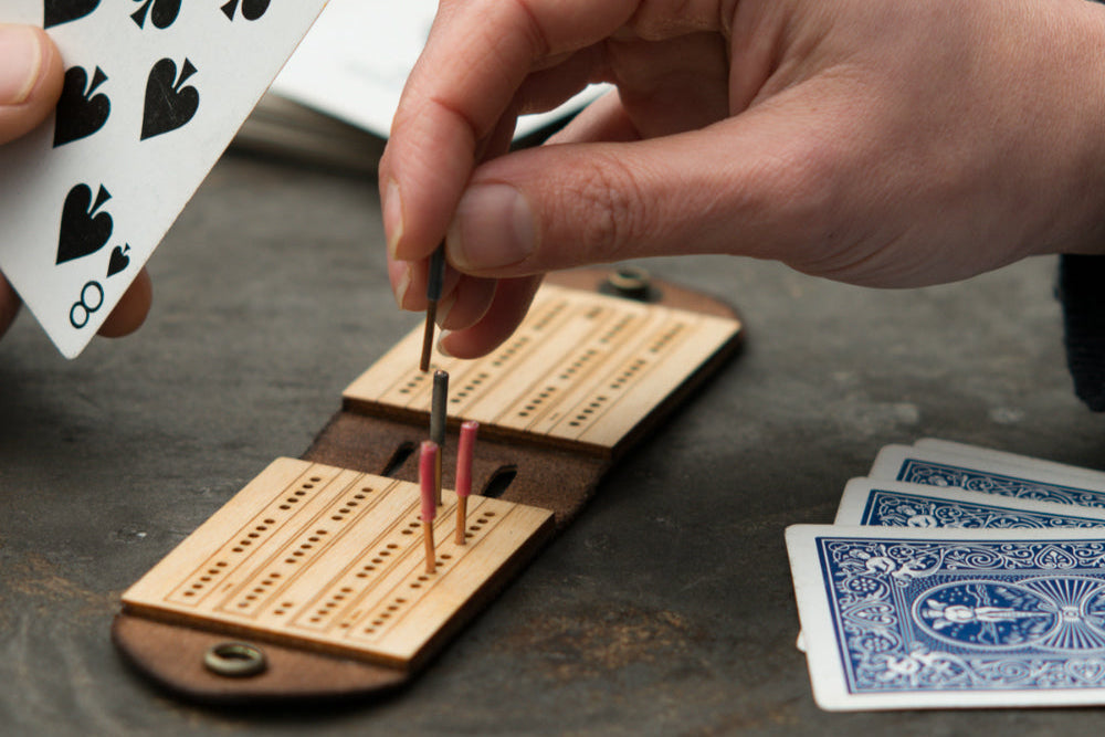 Two people playing a game of travel cribbage on a wooden picnic table with a small leather and birch cribbage board