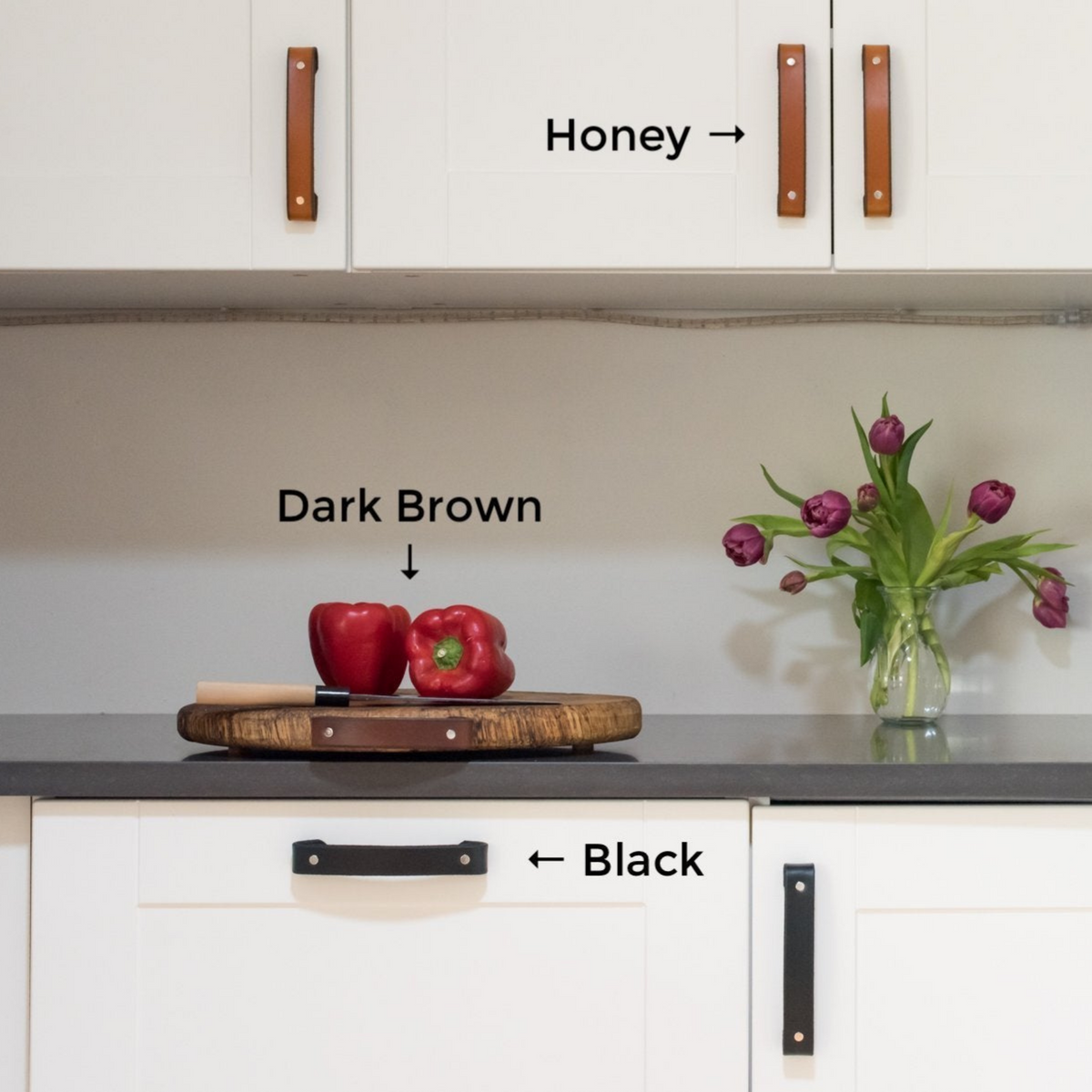 A white kitchen with gray countertops and 3 leather colors in one photo: Honey leather handles on the top cabinets, a dark brown leather handle attached to a wood cutting board, and black on the bottom cabinets. 