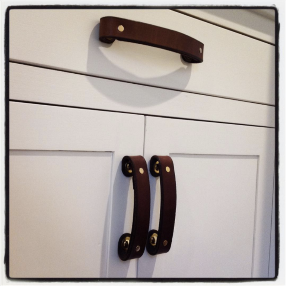 Customer photo showing dark brown leather handles with brass hardware on a white kitchen cabinet and drawer. 