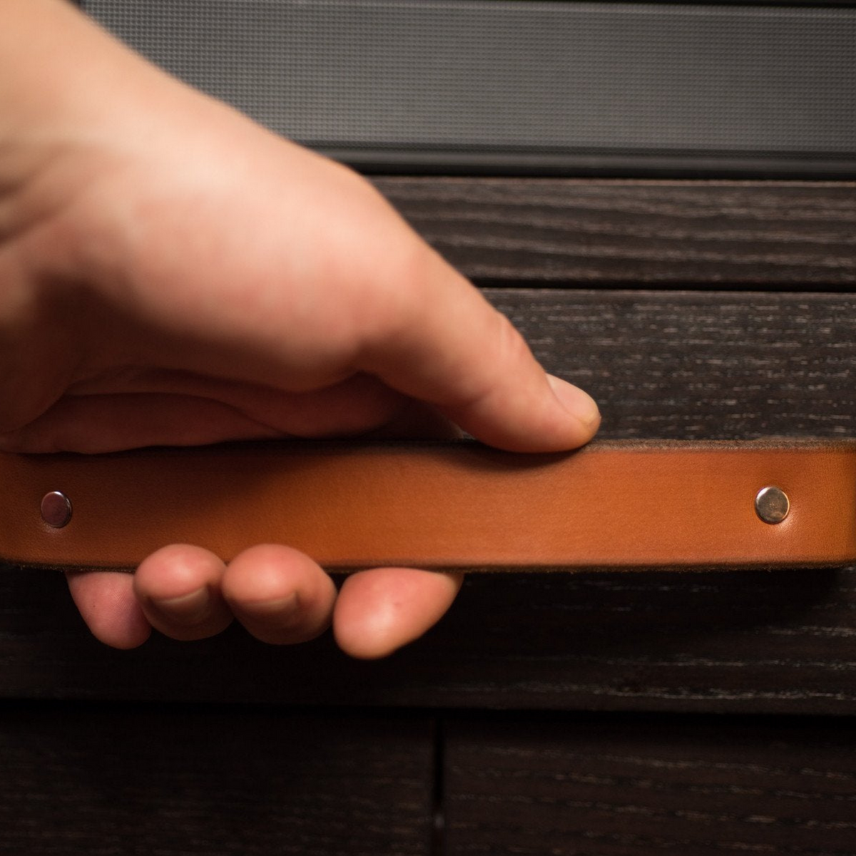 Close-up photo of a woman&#39;s hand in a large Tilikum leather handle. The handle is a honey / cognac color and the cabinet is a piece of IKEA furniture in black-brown.