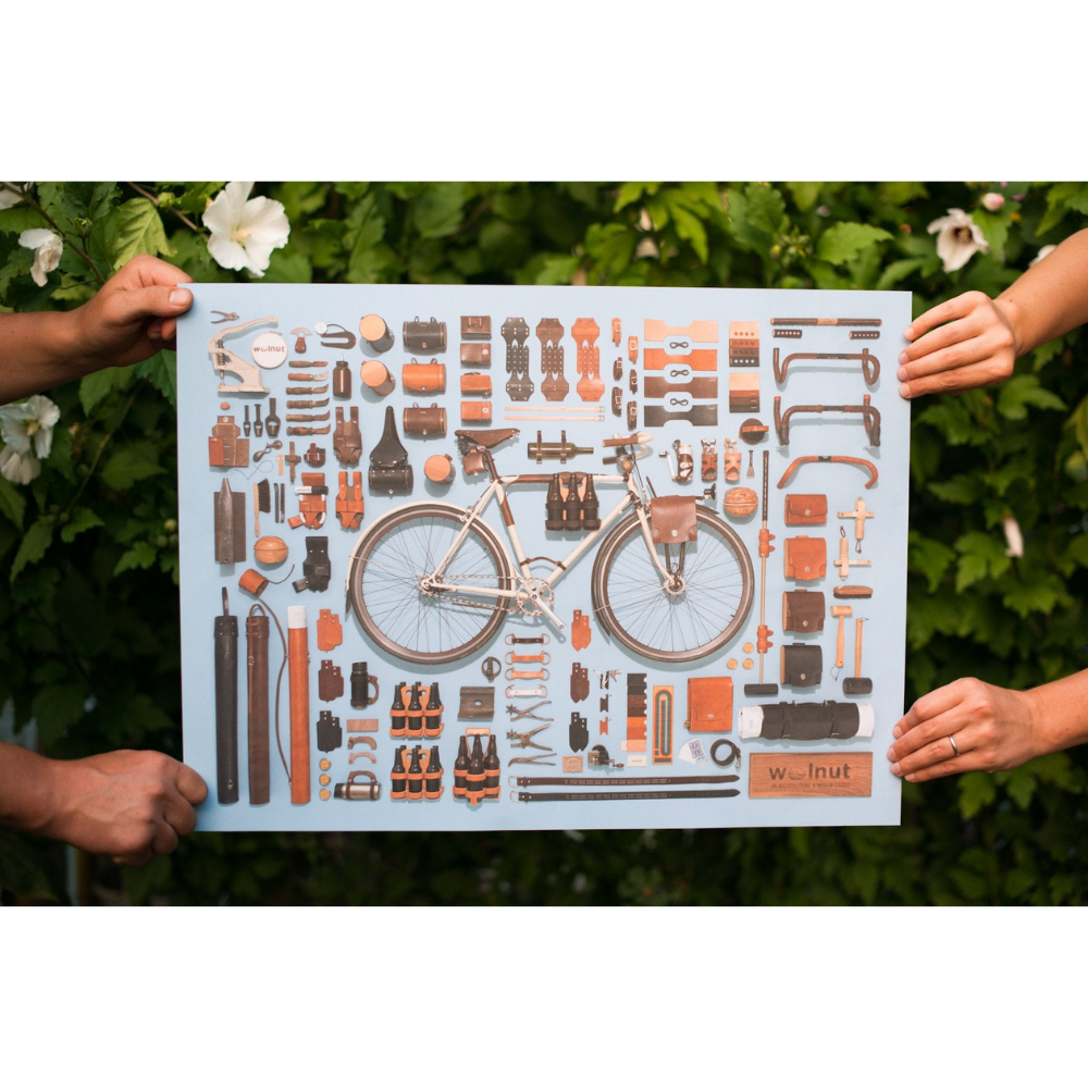 Four hands holding up an unframed bicycle poster with a variety of leather parts accessories