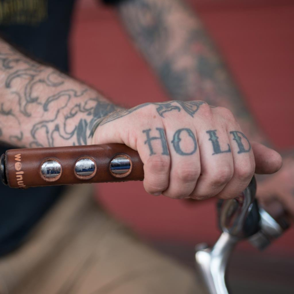 Tattooed man resting his hand on a bicycle city bar wrapped in a dark brown leather grip that has "porthole" round cutouts for finger holds. The man's hand tattoos say "HOLD FAST"