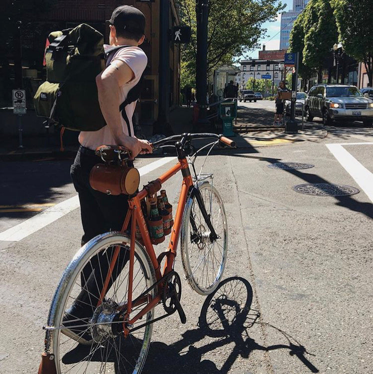 Customer photo of a bicycle being walked across a street intersection with a 6-Pack strapped to the top tube of the bicycle with a bicycle beer carrier combo in Portland Oregon