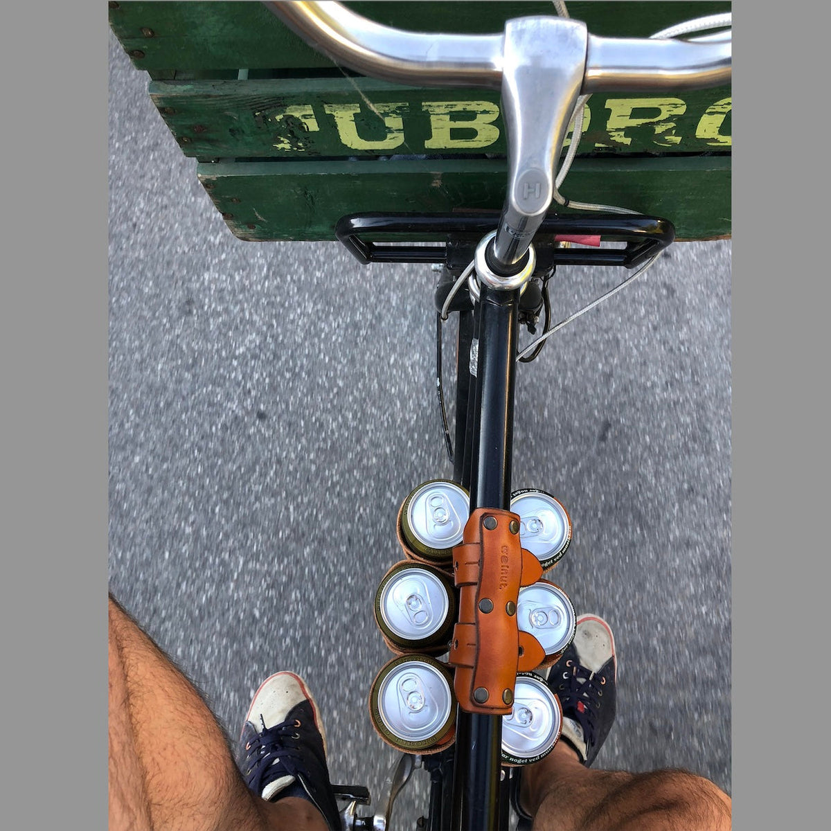 Customer photo looking down on the top tube of a bicycle from the saddle with their feet on the pedals. The street below is in motion and the top tube is holding a 6-pack of beer with a leather carrying strap