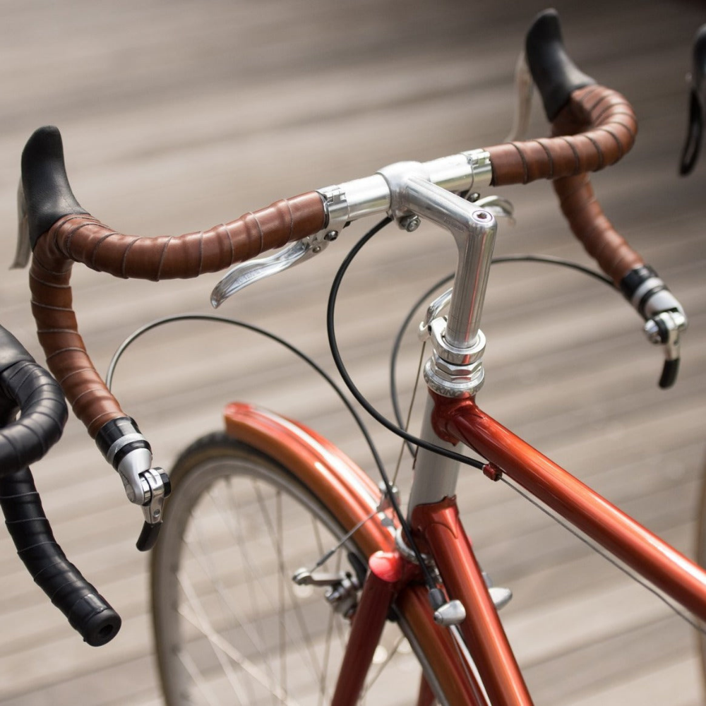 A bird&#39;s eye view looking down on three bicycles lined up in a row over a boardwalk floor. Each bicycle has a different color leather wrapped in coils on its handlebars: Dark Brown, Black and Honey color.