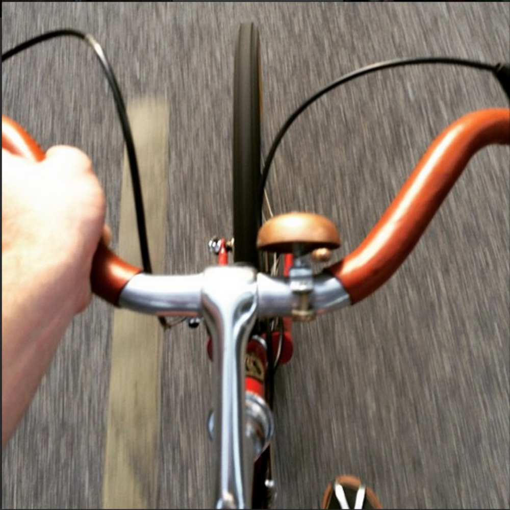 Photo taken by a customer on their bicycle from the point of the view of the saddle, as the rider. She is looking down towards her front tire and mustache handlebar, which is covered in honey colored sew-on leather bar wraps. The road below is blurred from motion.