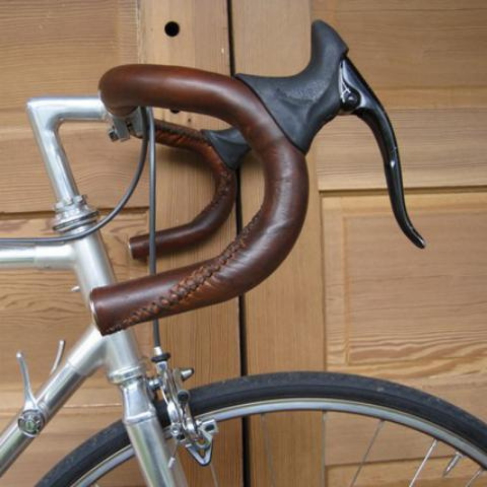 Dark Brown sew-on leather bar wraps on a set of bicycle handlebars  (drop bars). This is a close up profile view of the handlebars against a raw wood backdrop.