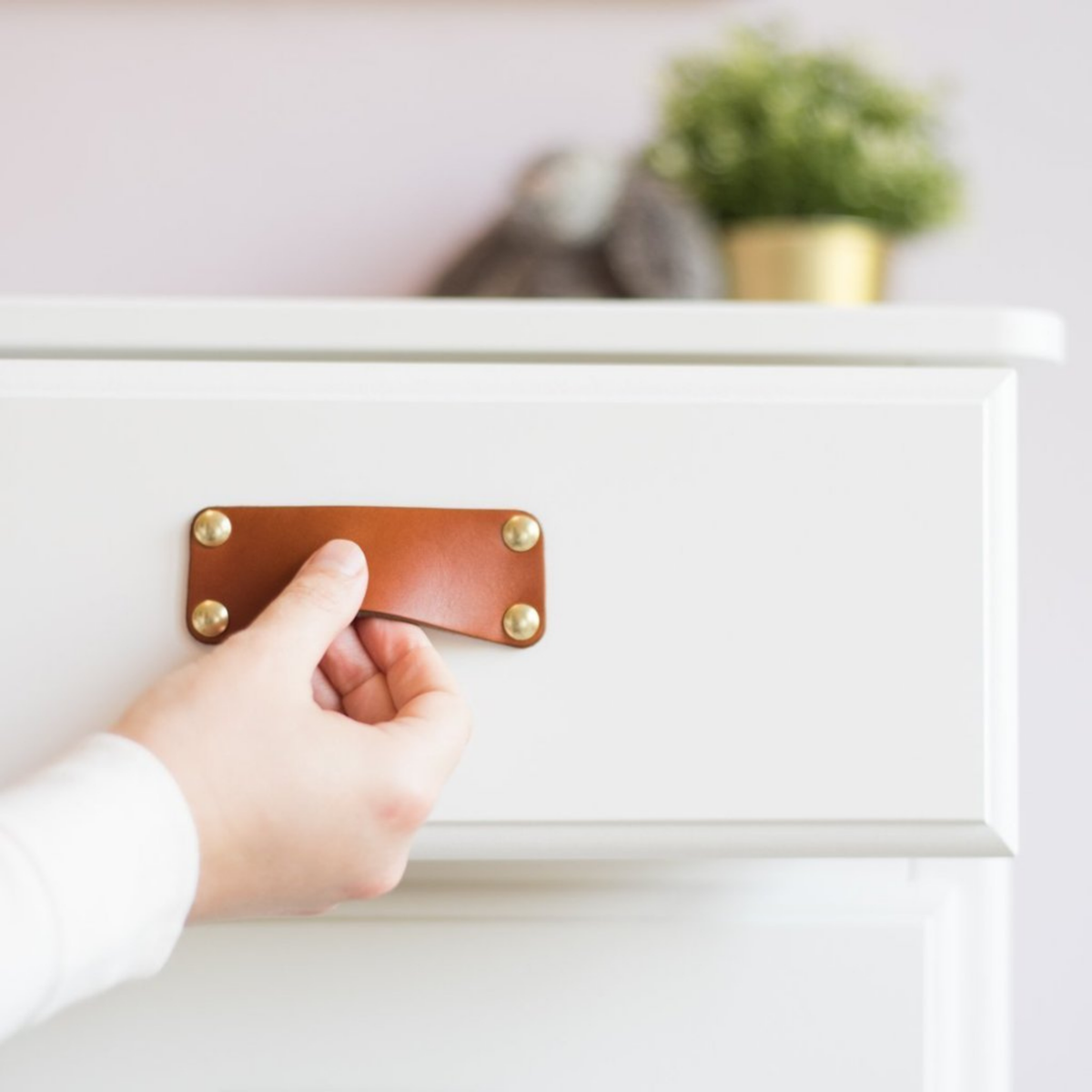 Woman&#39;s hand is opening a drawer in a white chest of drawers using a honey leather bin pull with brass hardware. 