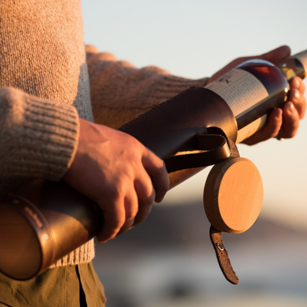 A man pulling a bottle of Scotch whiskey out of a hand-stitched leather case at sunset on the beach. The case's wood lid is flapped open but still attached to the case with a leather strap.