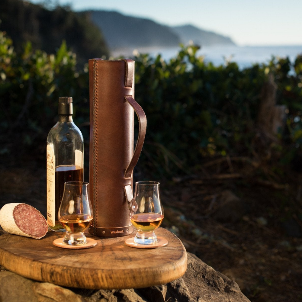 Still life scene of a leather whiskey bottle case, a bottle of Scotch, and two filled Glencairn glasses on a beautiful Oregon Coast beach with the blue sky in the background. 