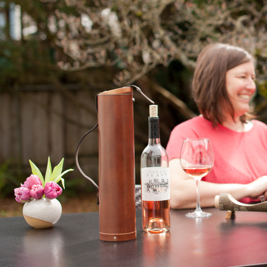 A smiling mother sitting at a table with a vase of fresh flowers, an open leather wine bottle case, an uncorked bottle of rose wine and a glass of wine.