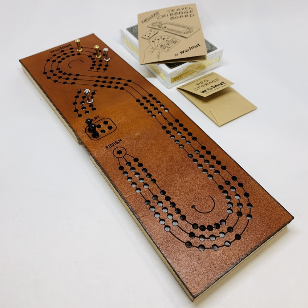 Unfolded Leather Travel Cribbage Board Laying Flat on Table Surface with All Associated Parts Displayed: 3 Sets of Pegs, Deck of Cards, Peg Storage Envelope with Mini Rules Booklet