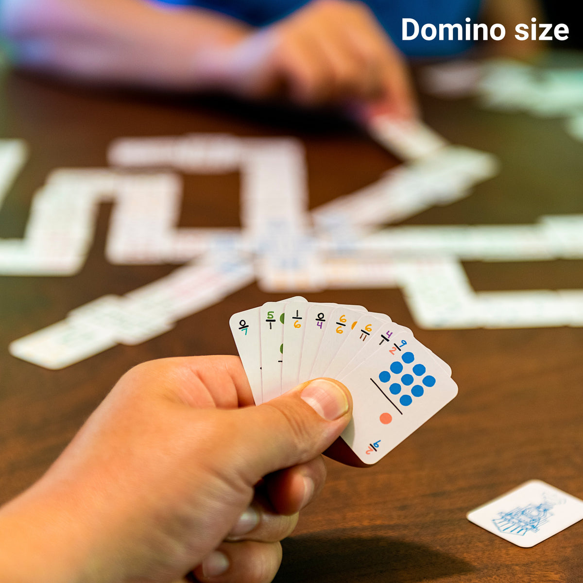 Travel dominoes in the smaller domino size on a table with two people playing. The cards are in one hand fanned out with index corner numbers visible. They are about the quarter of the size of a regular playing card. 
