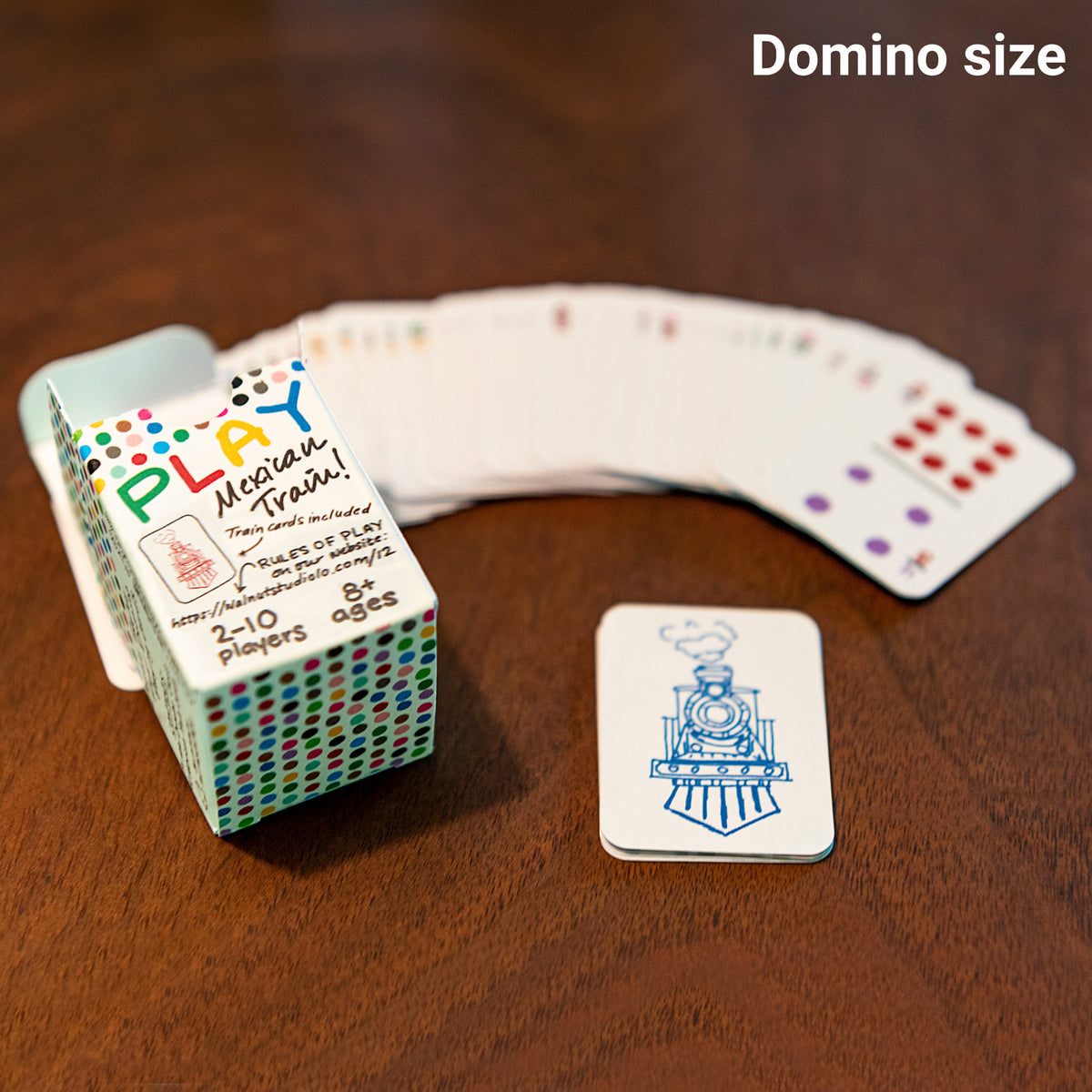 Close up of the box of domino-sized playing cards of double 12 dominoes . It is on the table in sharp focus along with a card showing a train for the Mexican Train game and the rest of the cards are fanned out in the background , blurred out.