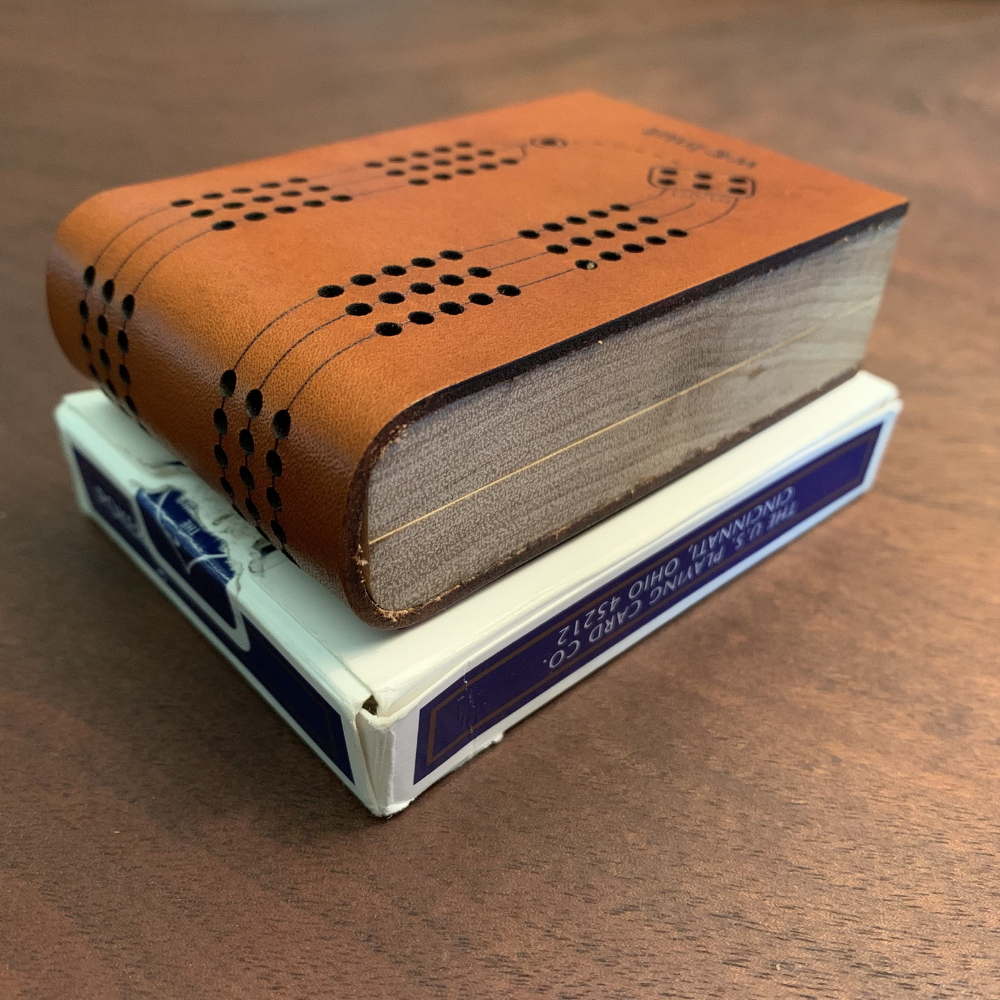 Shows closed Mini Deluxe Travel Cribbage Board on top of a regular deck of playing cards: ithe length and width is smaller than the cards but it is thicker.
