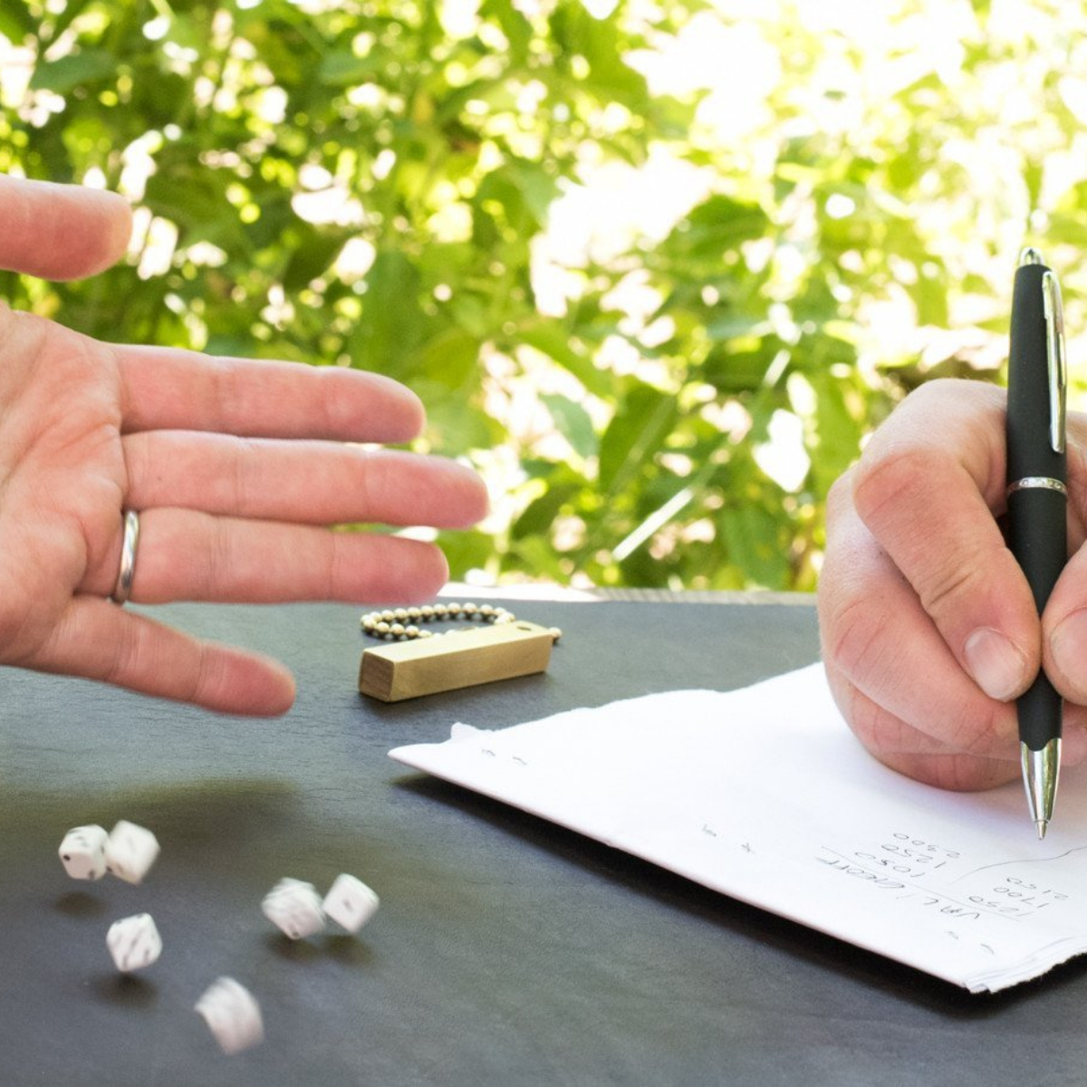 Two people playing dice on a summery day with green bushes in the background. One person is rolling six small travel dice and the other person is keeping score with a pen and paper. The brass storage tube is on the table.