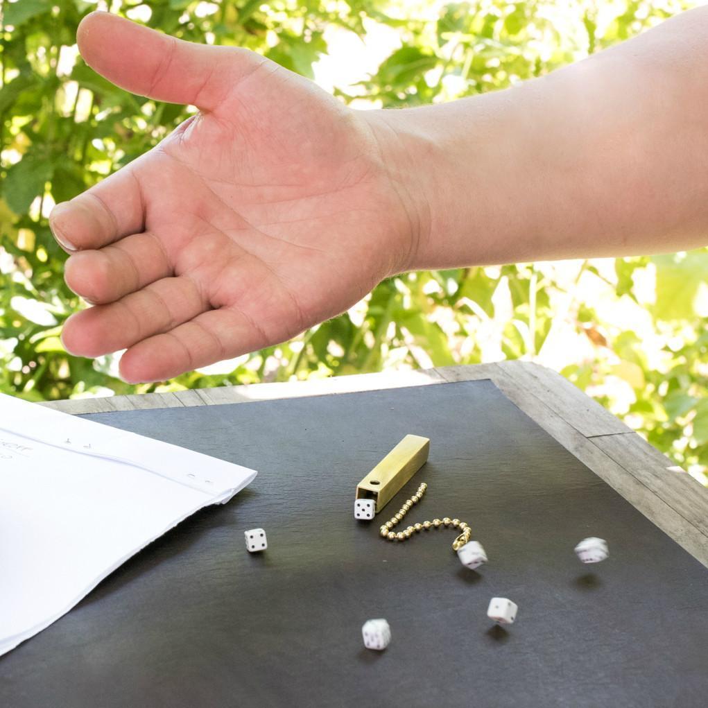 Man rolling six small white dice with black pips on a leather tabletop. On the table is the square brass tube that holds the dice and the brass beaded chain that serves as both a keychain and the closure for the tube.