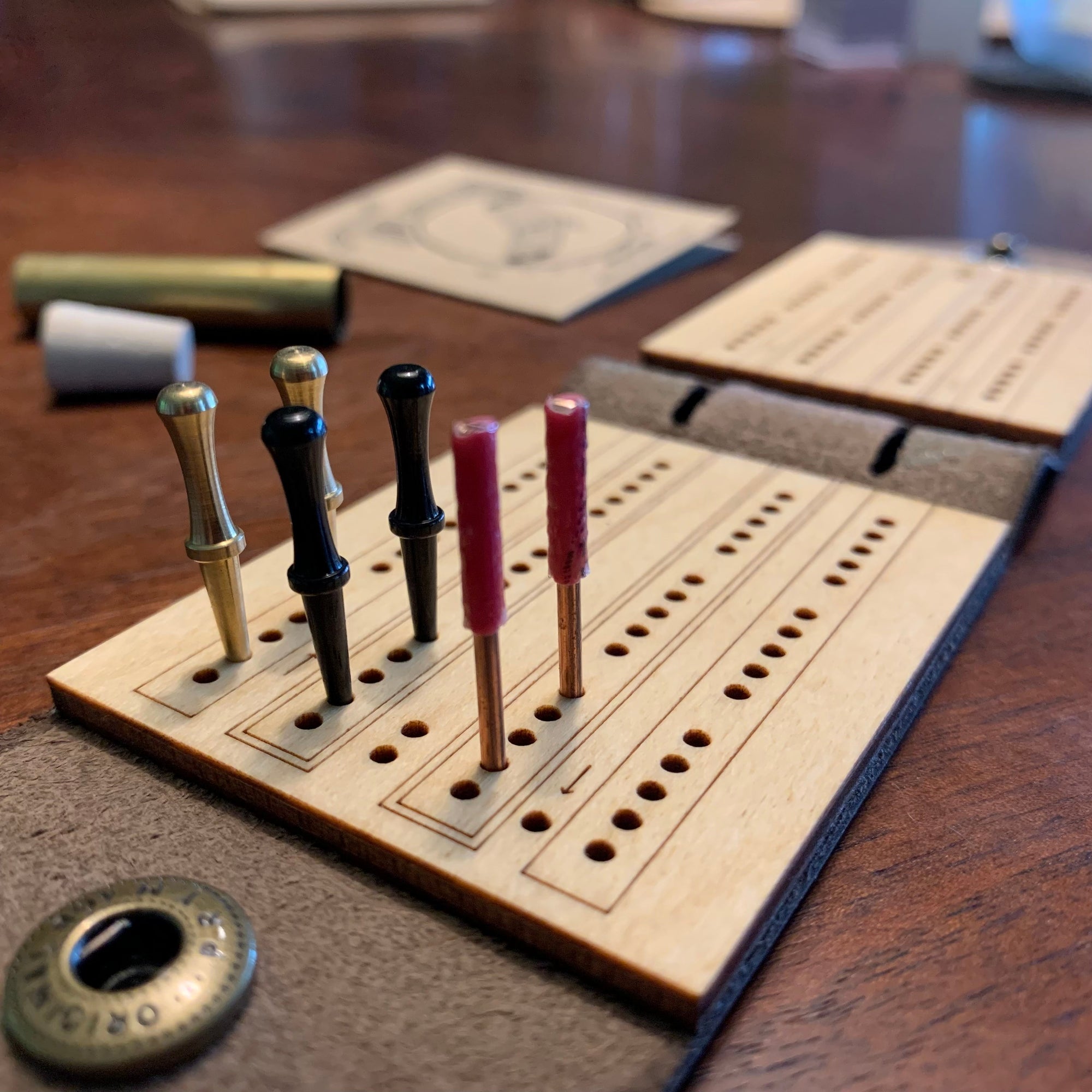 A small travel cribbage board is on a walnut wood table mid-game with 3 players card hands laid out and a third set of pegs parker on the 2nd track going backwards