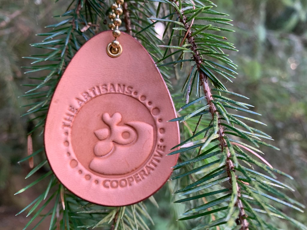 A custom egg-shaped leather ornament embossed with the ARtisans Cooperative corporate logo hanging from a blue spruce tree branch