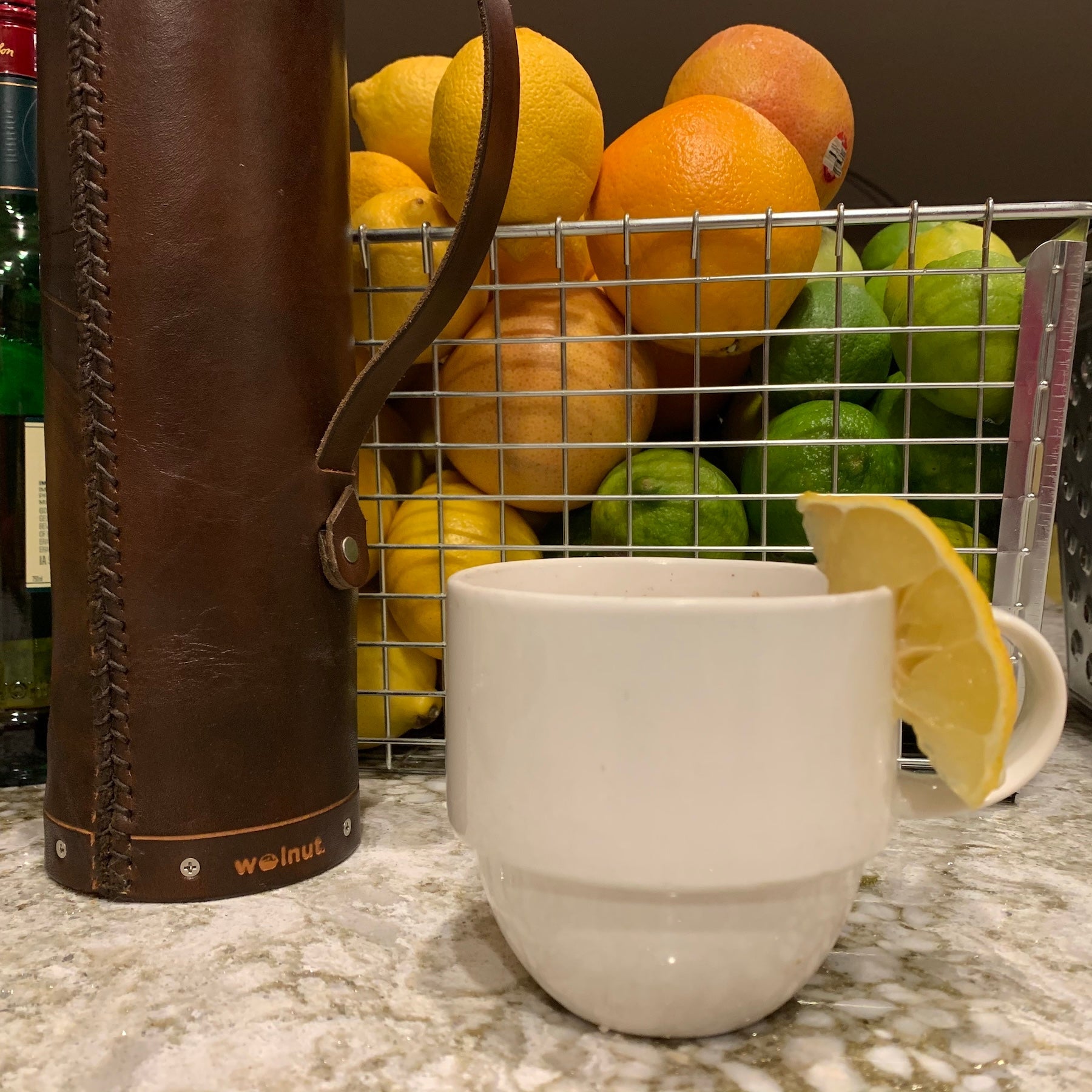 A white mug with a lemon wedge on the rim sitting in front of a wire basket full of different citrus (orange, lemon, lime) and handmade leather whisky tube case