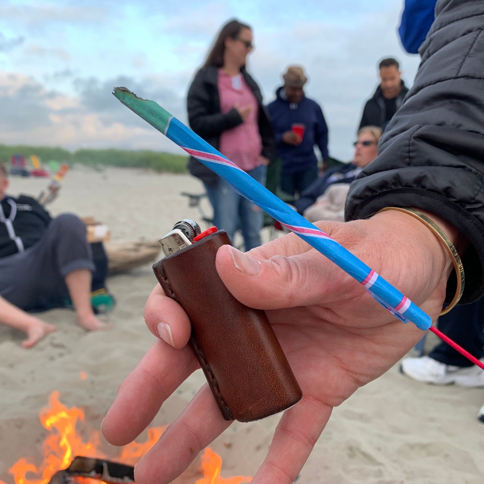 A woman's hand holding a sparkler on the 4th of July at a party on a public beach with a lighter in a handcrafted leather lighter sleeve 