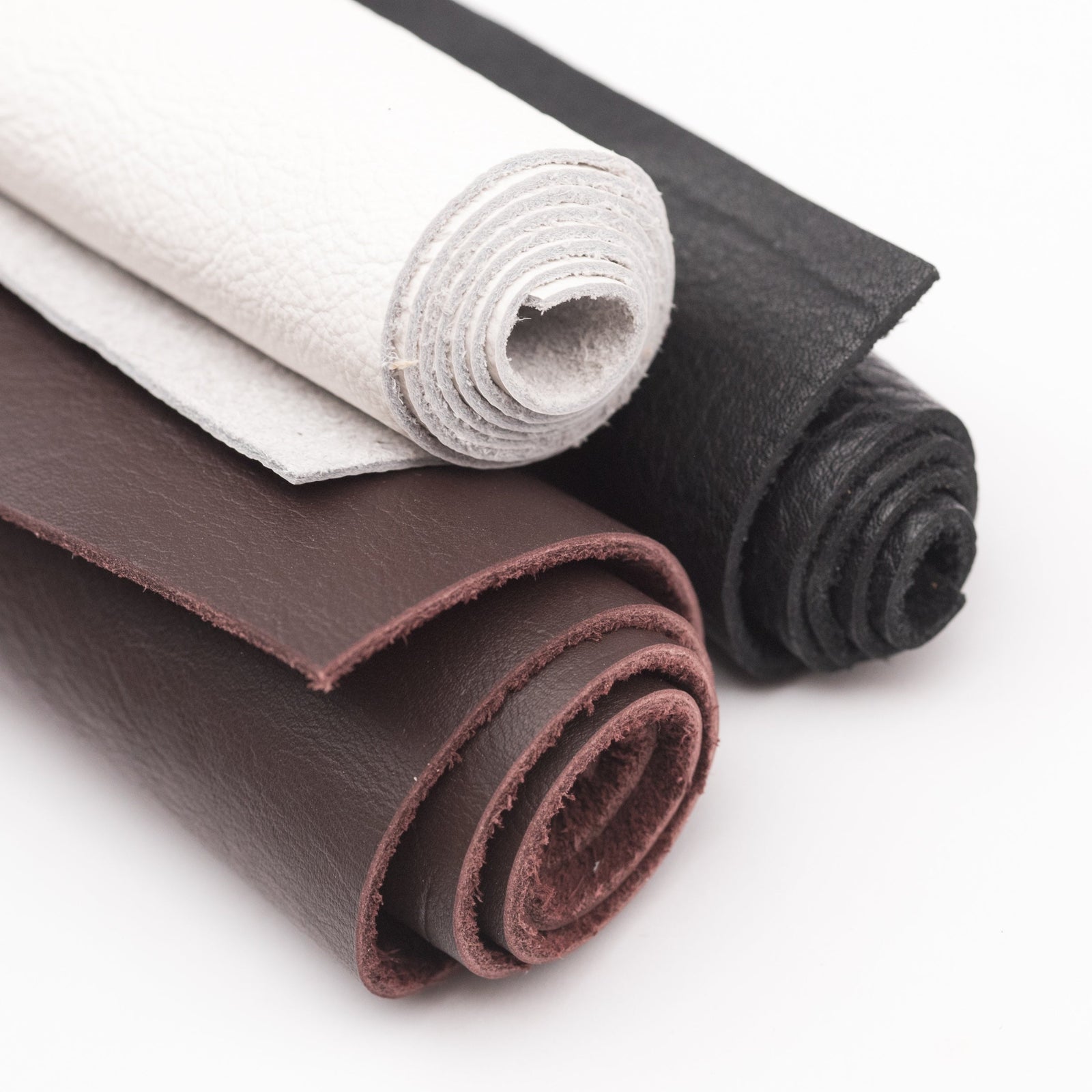 Three rolls of leather on a white background: white garment-tanned, dark brown bridle tanned, and black top grain