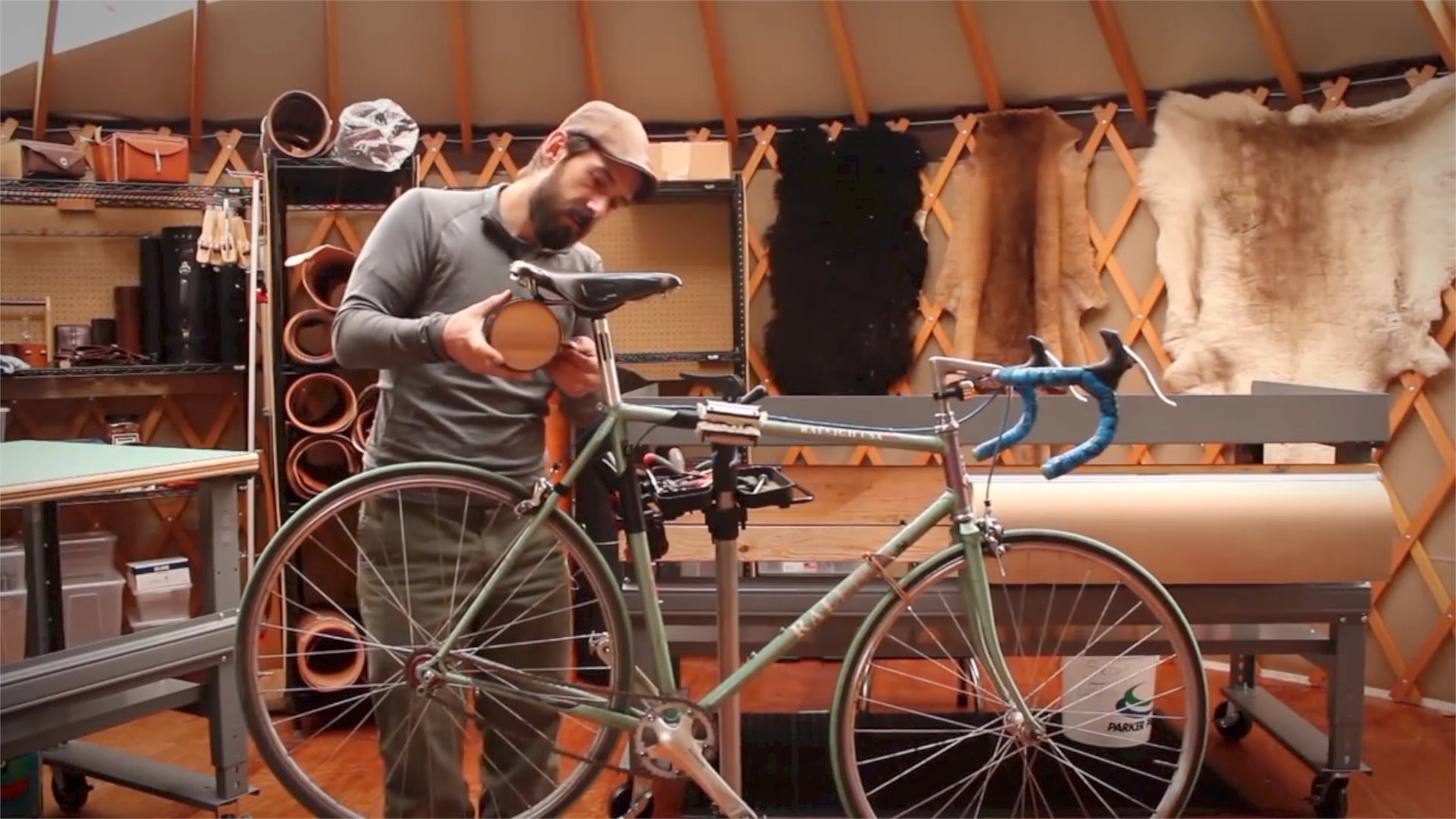 Geoff Franklin of Walnut Studiolo installing a leather bicycle barrel bag on the saddle of a bicycle in a yurt, surrounded by hanging sheepskins and rolls of leather in warm lighting
