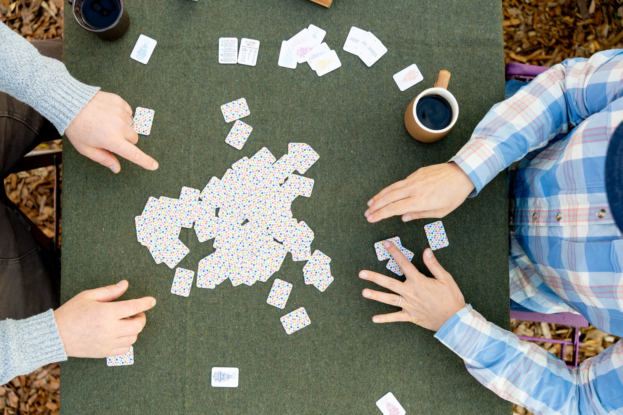 Bird's eye view of a table while two people are playing domino and picking their hands from the pile of tiles or boneyard, using the new version 2 of the double 12 travel domino playing cards. The table has a green wool background and they are drinking cu