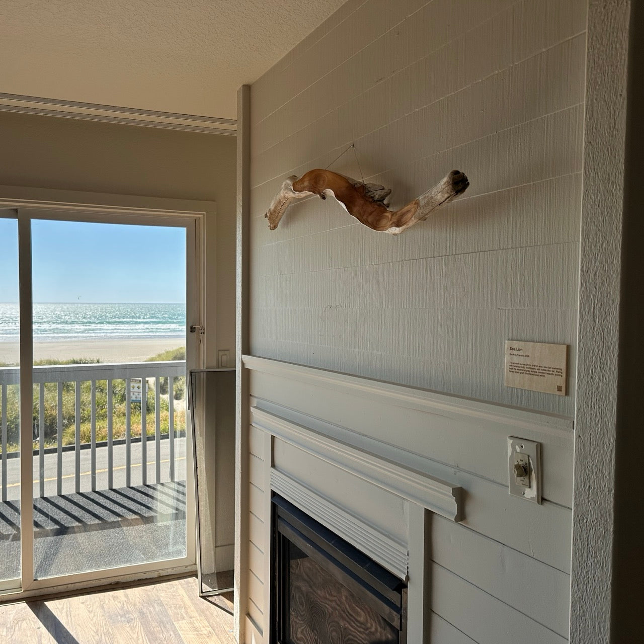Looking out a hotel room towards a sliding glass door with views of the beach and ocean on a sunny day, and a driftwood sculpture hanging over the fireplace mantel to the right