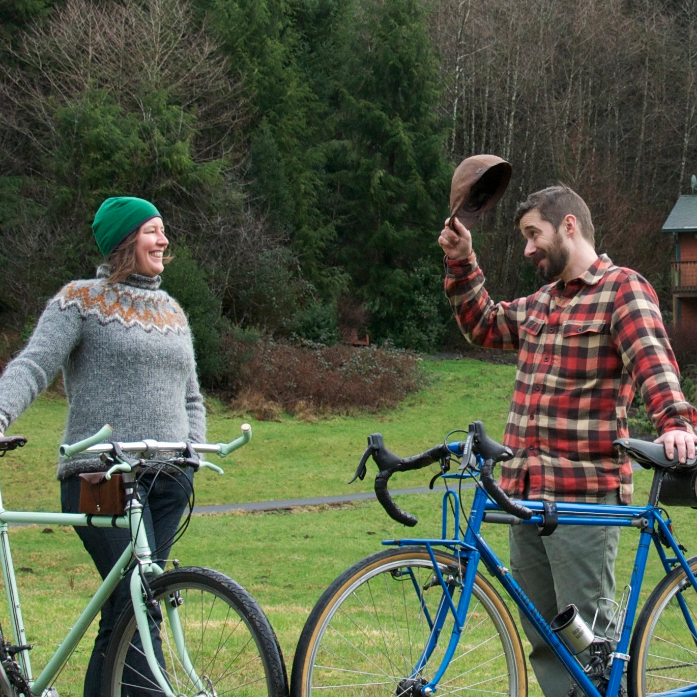 Geoff tips his hat to Valerie in a gesture of thanks, appreciation and gratitude while they each stand with bikes on a green Oregon meadow surrounded by Doug Fir trees