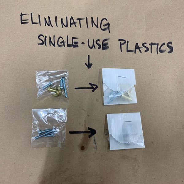 Handwriting of black marker on a Kraft paper background that says, Eliminating Single Use Plastics, with two sets of hardware kits and directional arrows below showing the packaging changing from plastic to paper