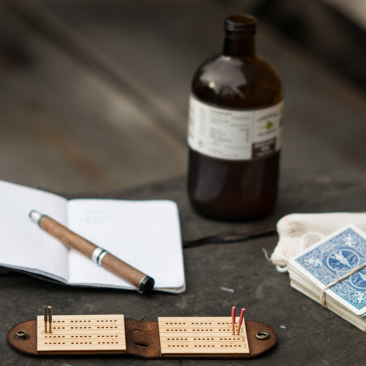 A small cribbage board on a gray wood table with a notebook, deck of cards, and a bottle of kombucha
