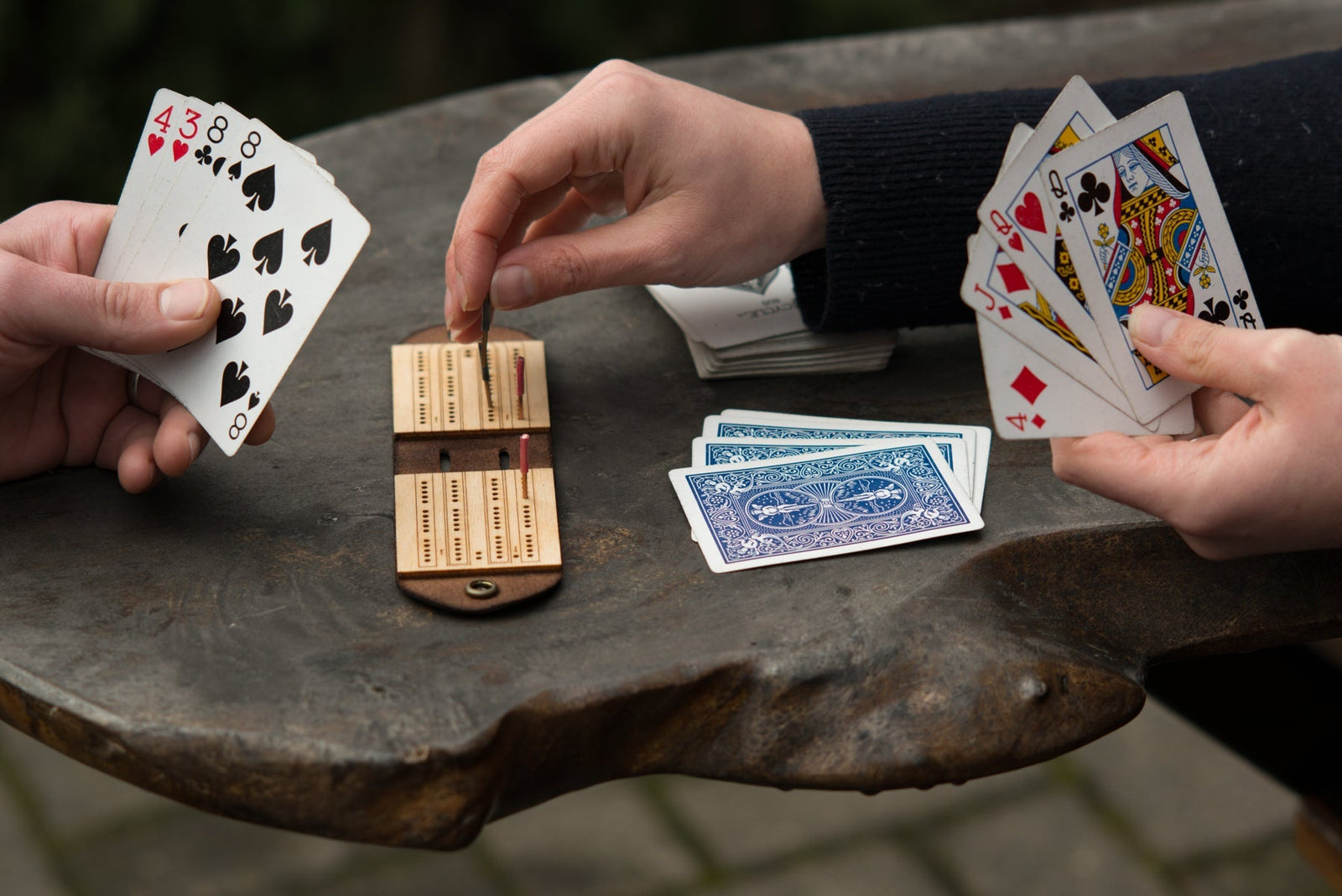 Two people are playing cribbage with a small leather and wood travel-sized game board on a picnic table