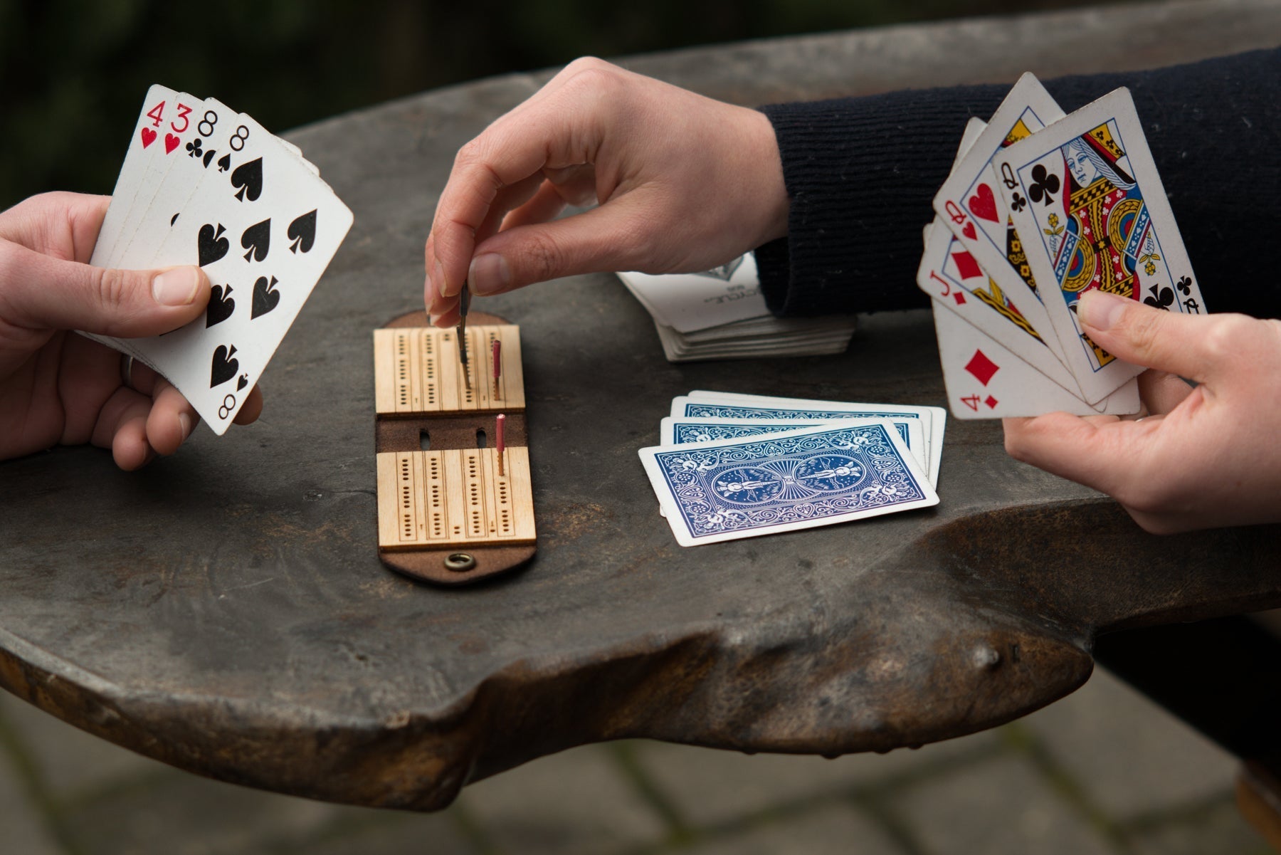 Two people are playing cribbage with a small leather and wood travel-sized game board on a picnic table
