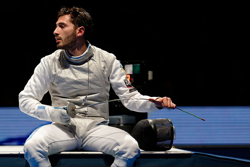 ﻿﻿A young Italian man sitting sweaty in an Olympic stadium, wearing a full white protective fencing outfit and wiping his foil fencing blade