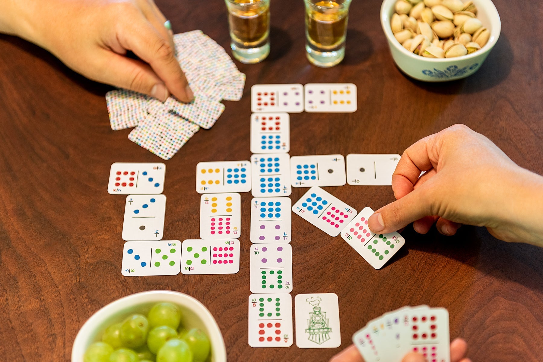 Two people are playing dominoes using colorful hand-drawn unique playing cards dominoes on a beautiful walnut wood surface with travel snacks on the table, pistachios and grapes and a shot of whiskey